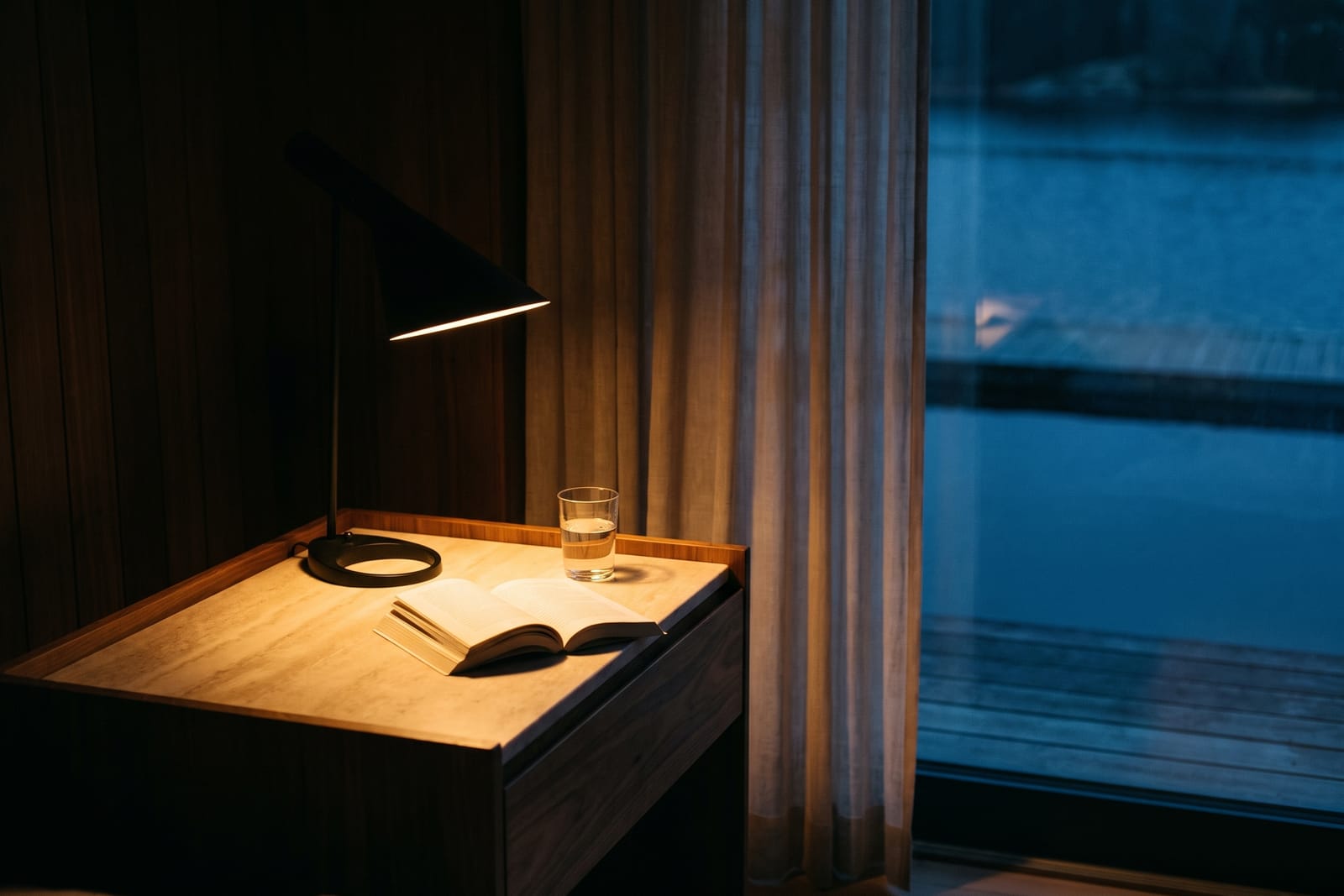 Bedside table late at night with a lit reading lamp, an open book face down and a glass of water in a dark, quiet bedroom