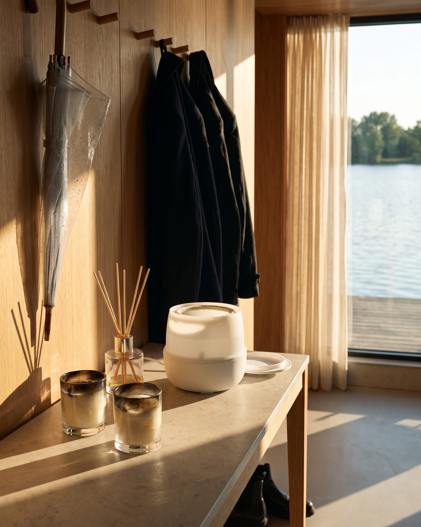 Hall console with two jar candles burnt down to a black ring, a reed diffuser and a plain white water mister, coats on pegs and a wet umbrella behind