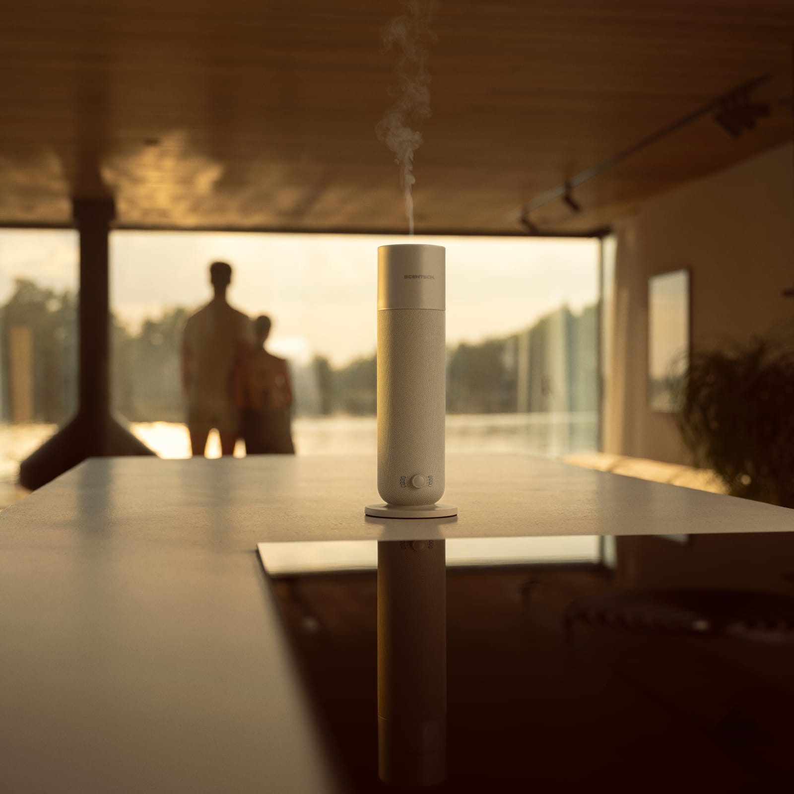 Smart Aroma Diffuser 2 releasing a fine mist on a long table at sunset, two people by the window in the distance